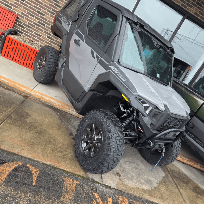 A gray all-terrain vehicle with large tires sits on a wet concrete surface in front of a brick building with large windows.