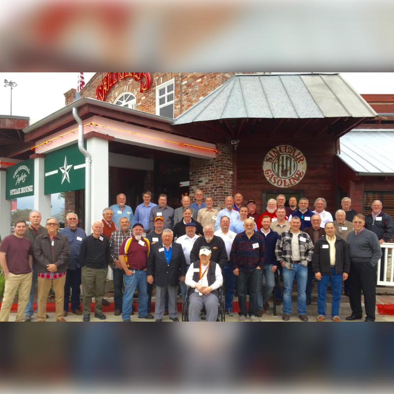 A group of men in casual attire pose for a photo in front of a brick building with a sign that reads "Sinclair Gasoline."