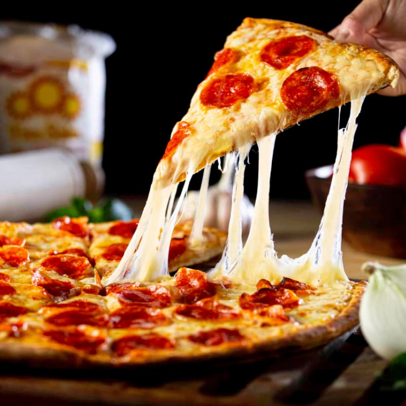 A hand lifts a cheesy slice of pepperoni pizza from a whole pie, with ingredients like tomatoes and onions visible in the blurred background.