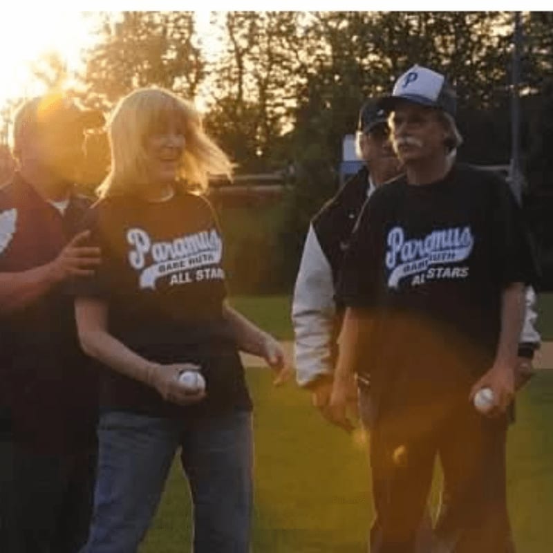 Four people in baseball attire stand on a field with trees and a hazy sunset in the background.