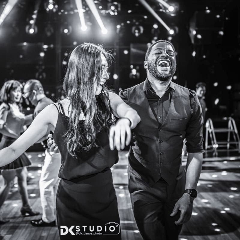 A joyful couple dances in the foreground while a disco ball illuminates the background with sparkling lights.