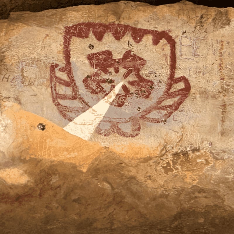 A red, shield-like petroglyph with intricate designs is painted on a rough, textured rock face, with faint scratches and markings visible in the background.