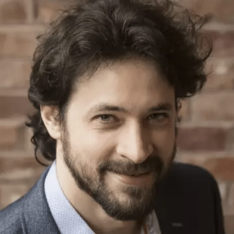 A man with dark, curly hair and a beard smiles at the camera in front of a brick wall.