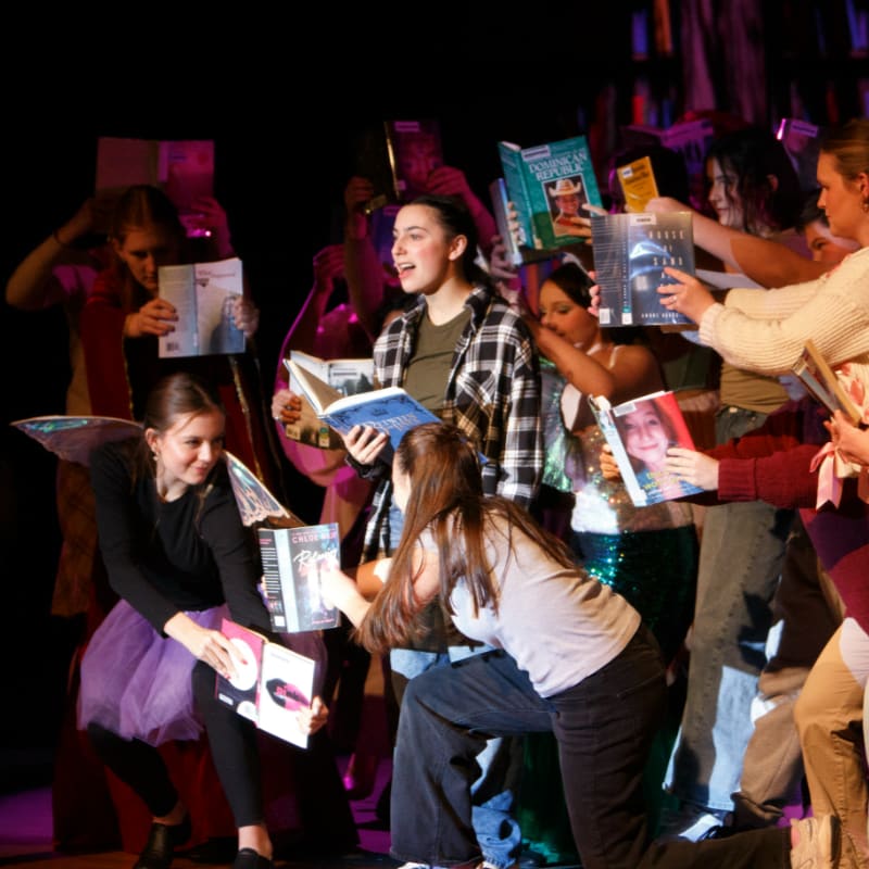A group of young people hold up various books and magazines in the foreground against a dark, indistinct background.
