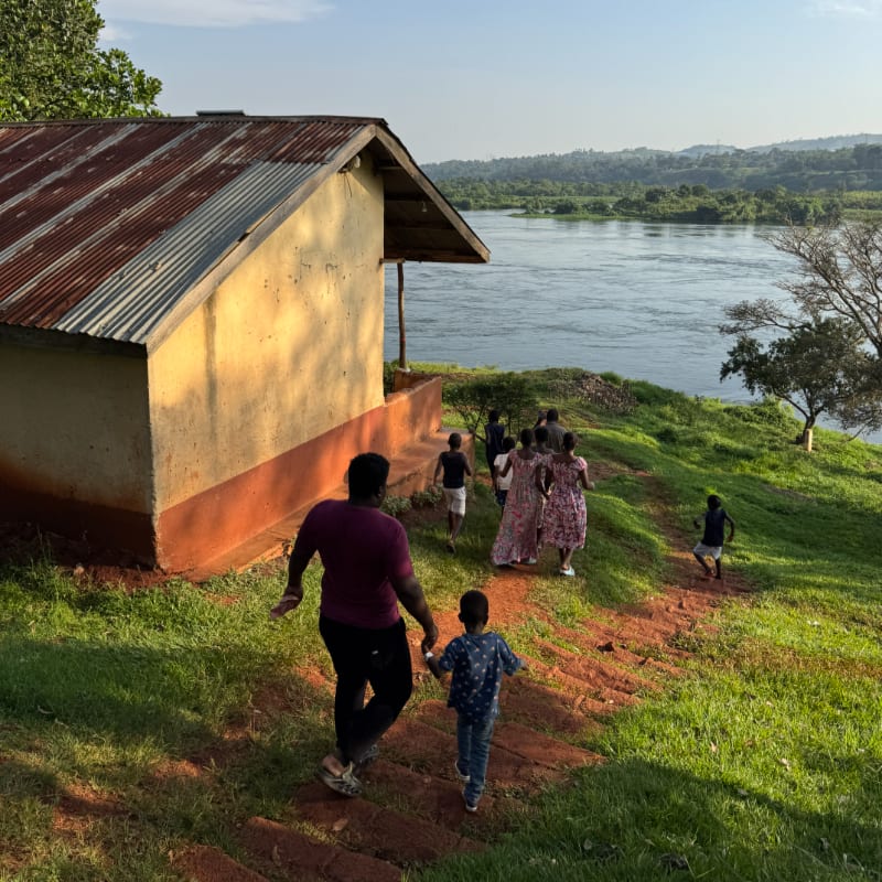 A man and a young child walk up a grassy path toward a group of people near a small house, with a wide river and distant hills in the background.