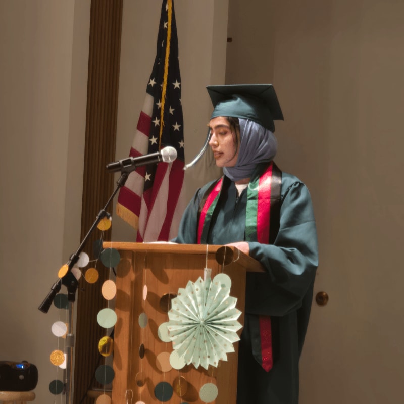 A graduate in a green gown and hijab speaks at a decorated podium with an American flag in the background.