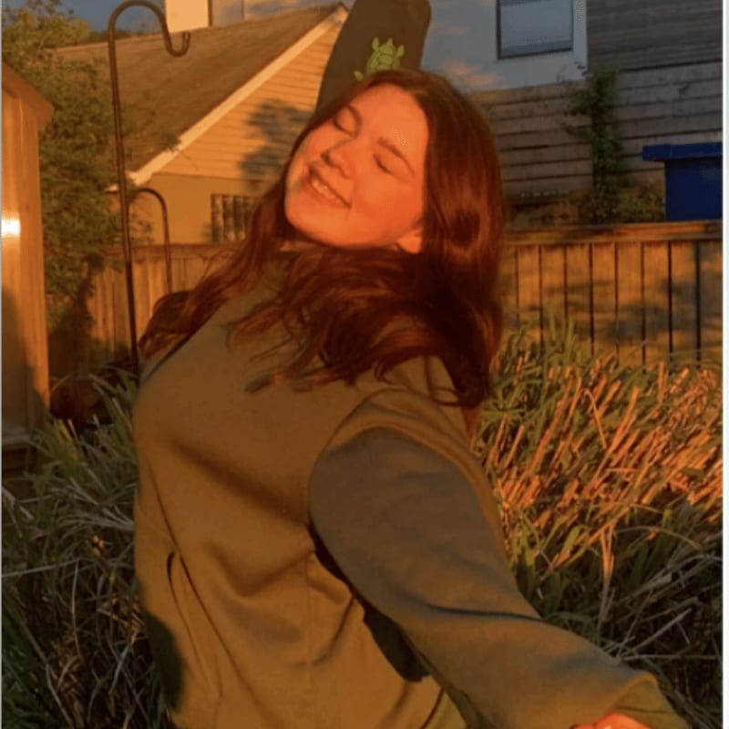 A smiling woman in an olive green sweatshirt stands in a garden with tall grass and a wooden fence and house in the background.