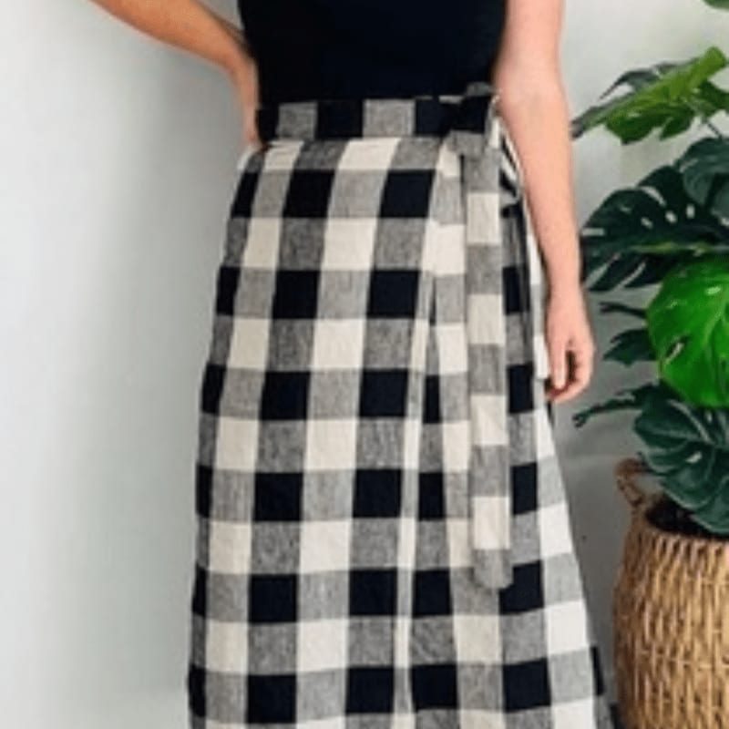A person wears a black and white checkered wrap skirt against a plain white wall and a potted Monstera plant.