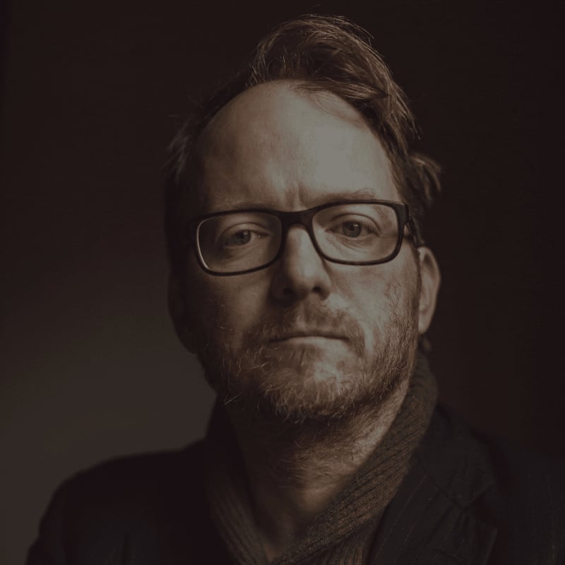 A man with glasses and a beard stands in the foreground against a dark, blurred background.