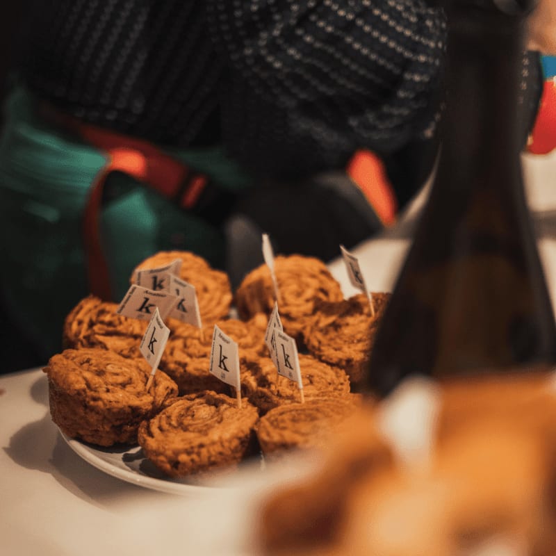 A plate of muffins with small flags sits on a table in front of a blurred bottle and a person wearing an apron.