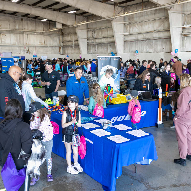 People gather around blue tables in a large warehouse, with a crowd of attendees and event displays filling the background.
