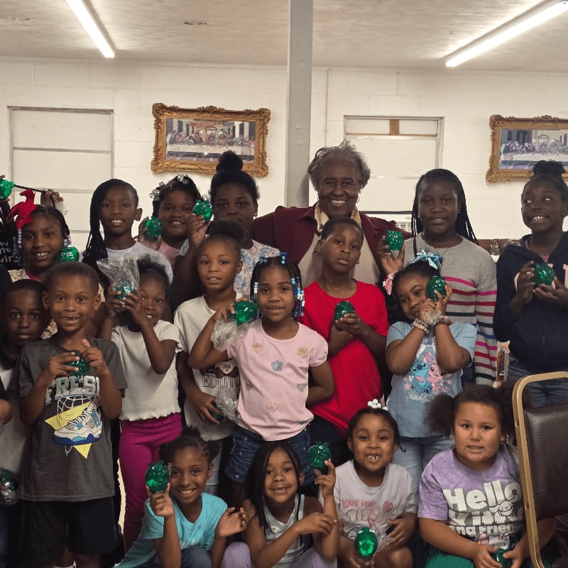 A group of children and an adult woman hold green ornaments in a room with white walls and framed pictures.