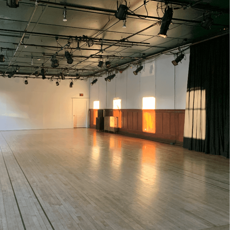 Sunlight streams through windows onto a polished wooden floor in an empty studio space with theatrical lighting overhead.