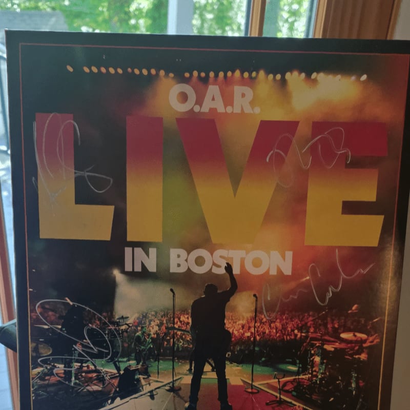 A signed O.A.R. "Live in Boston" concert poster featuring a silhouette of a performer facing a crowd is positioned against a blurred background of a window and greenery.