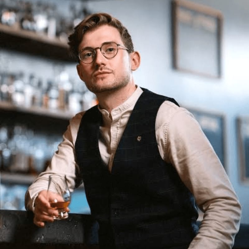 A man wearing glasses and a dark vest holds a glass of liquor in the foreground, set against a blurred bar background with shelves of bottles and framed art.