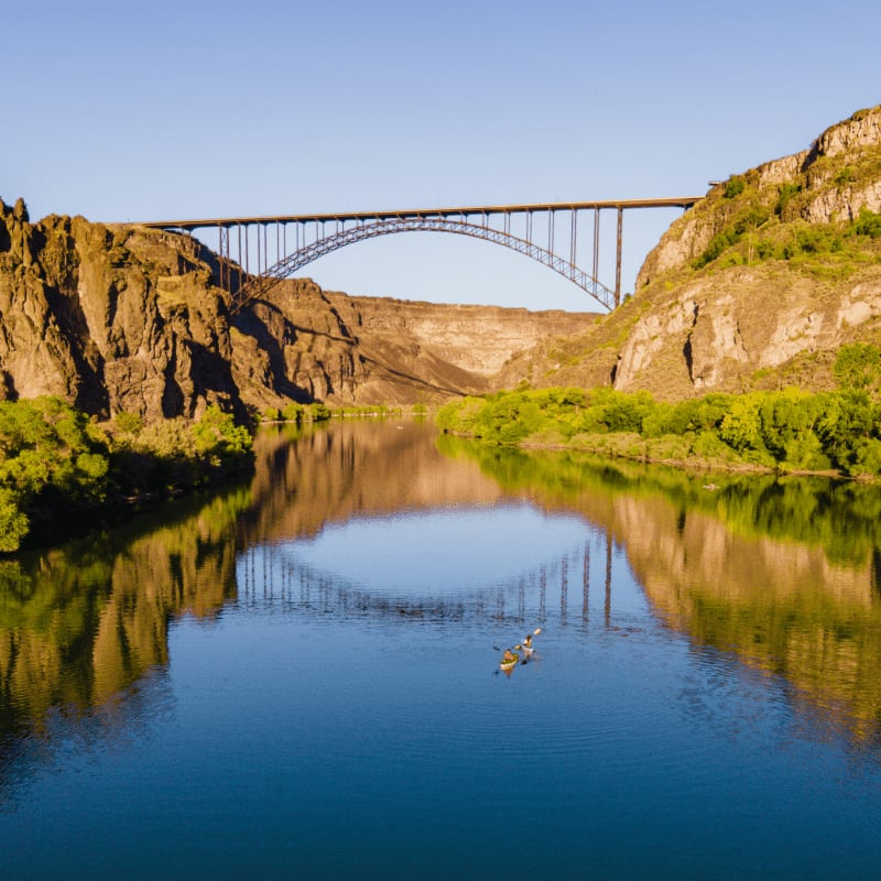 A lone kayaker paddles through a calm river in the foreground, reflecting a massive arched bridge and rugged canyon walls in the background.