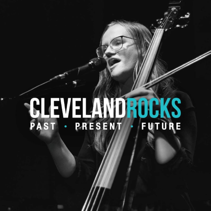 A woman with glasses sings into a microphone while playing a double bass against a dark, blurred background, with "CLEVELAND ROCKS" text overlaid in the center.