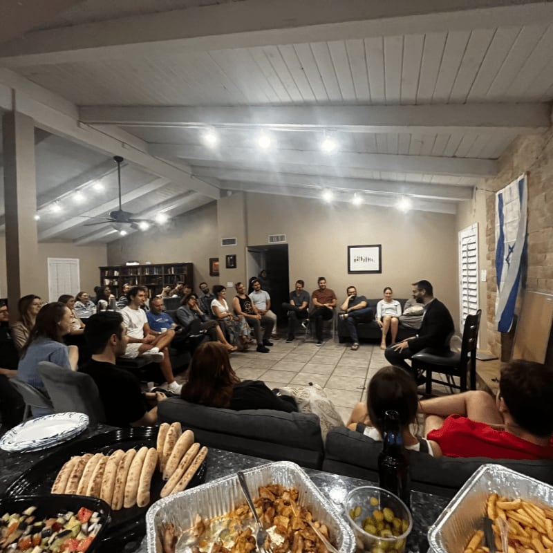 Platters of food sit in the foreground of a room where a group of people are gathered in a circle for a discussion.