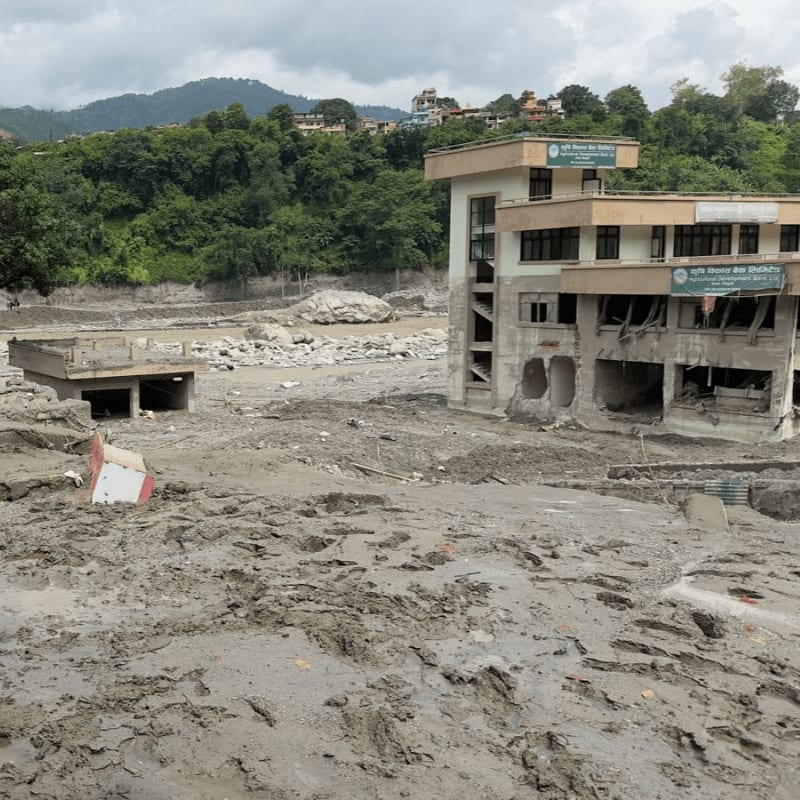 A damaged multi-story building stands amidst a landscape of mud and debris in the foreground, with a lush green hillside and a cloudy sky in the background.