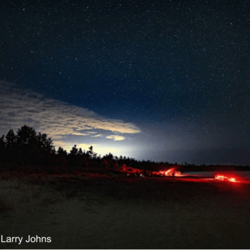 Glowing red fires burn on a dark landscape under a vast, starry night sky with a sliver of clouds.