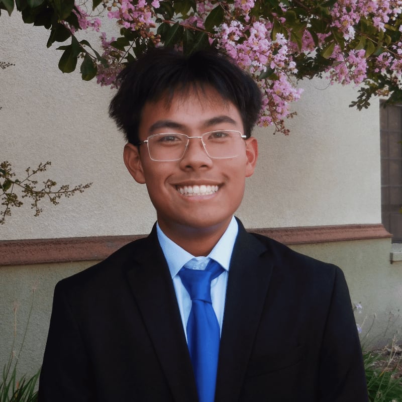 A smiling young man in a black suit and blue tie stands before a neutral-colored wall topped with pink blossoms.