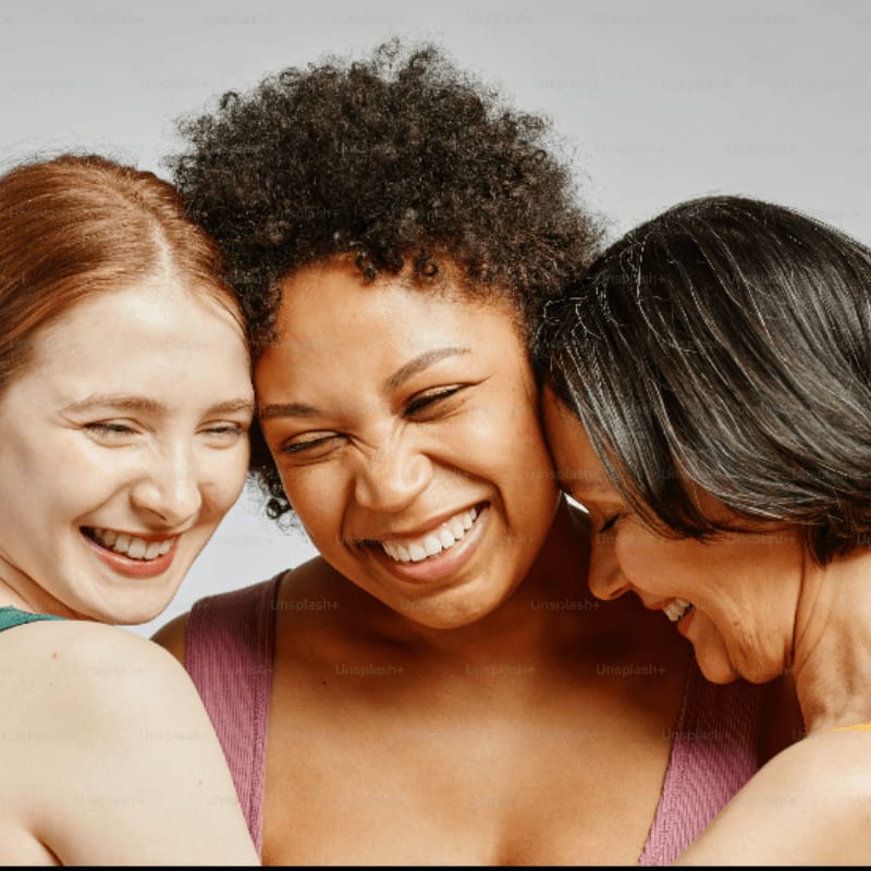 Three smiling women of different ethnicities huddle closely together against a plain, light gray background.