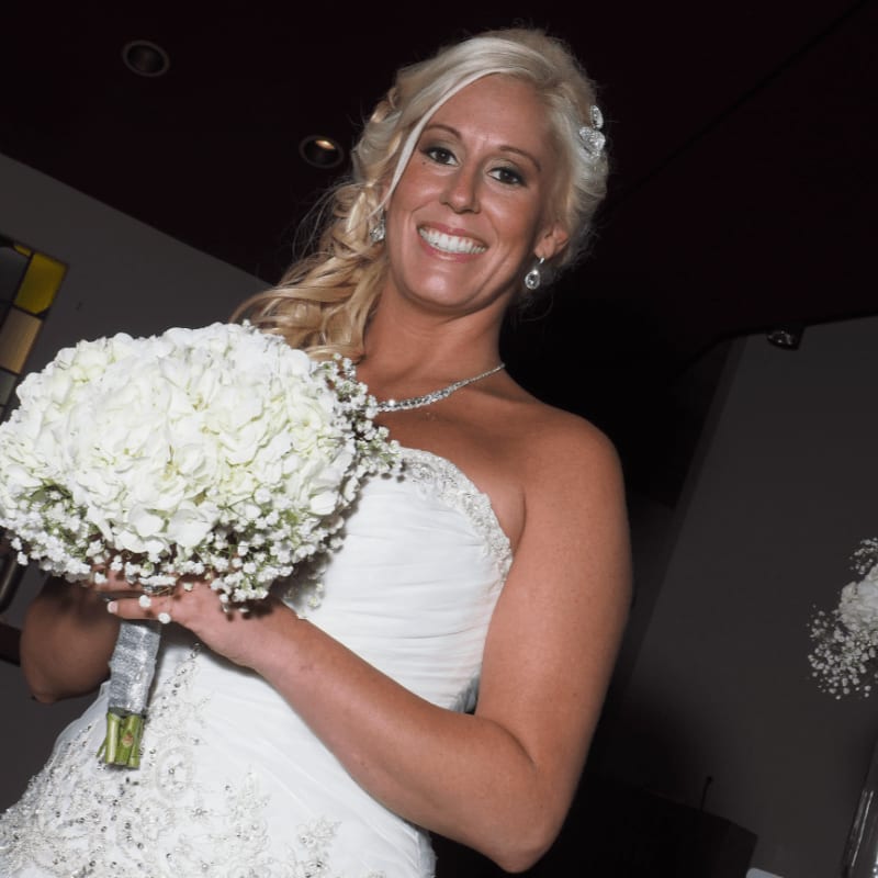 A smiling blonde bride in a white gown holds a large white bouquet against a dark, dimly lit background.