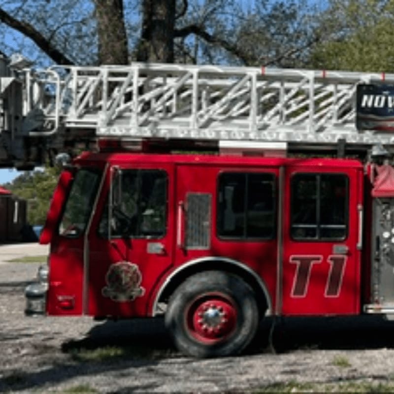 A red fire truck with a large white ladder on top is parked on a gravel surface with trees in the background.
