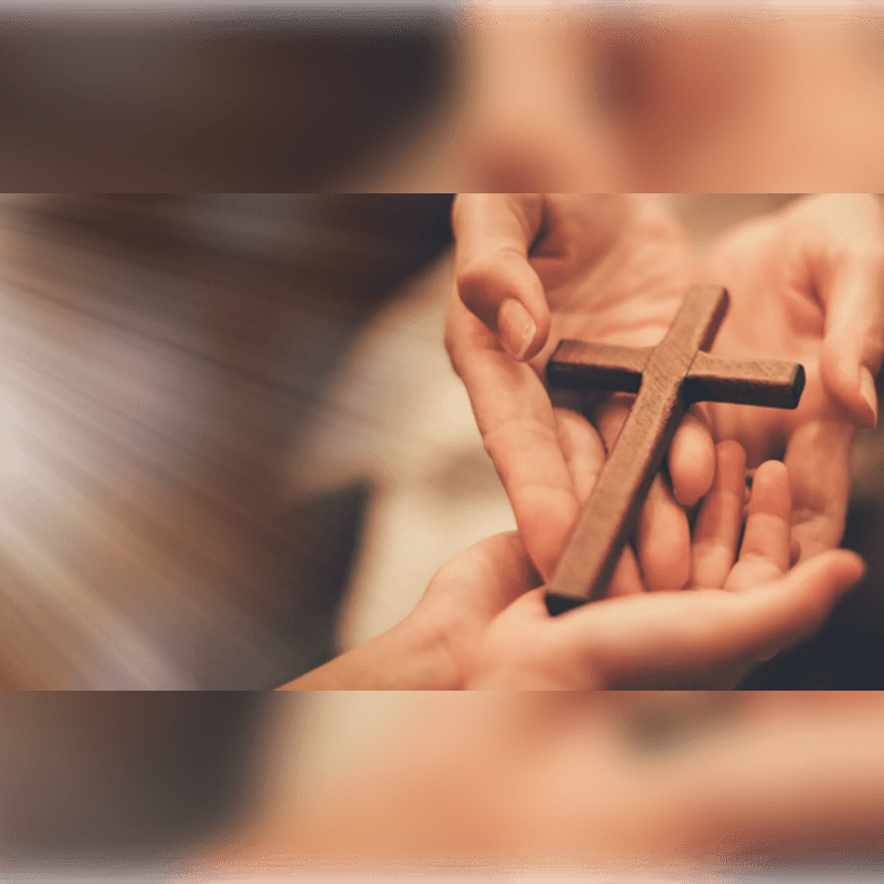 Two hands gently cradle a small wooden cross against a soft, blurred background.
