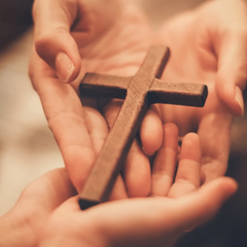 A small wooden cross is cradled in a pair of open hands against a soft, blurred background.
