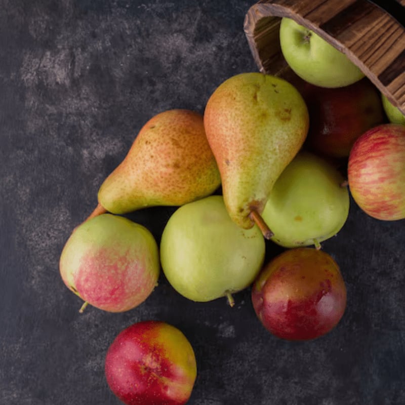 A variety of red and green apples and pears are scattered across a dark, textured surface, with some spilling out of a wooden bucket in the background.