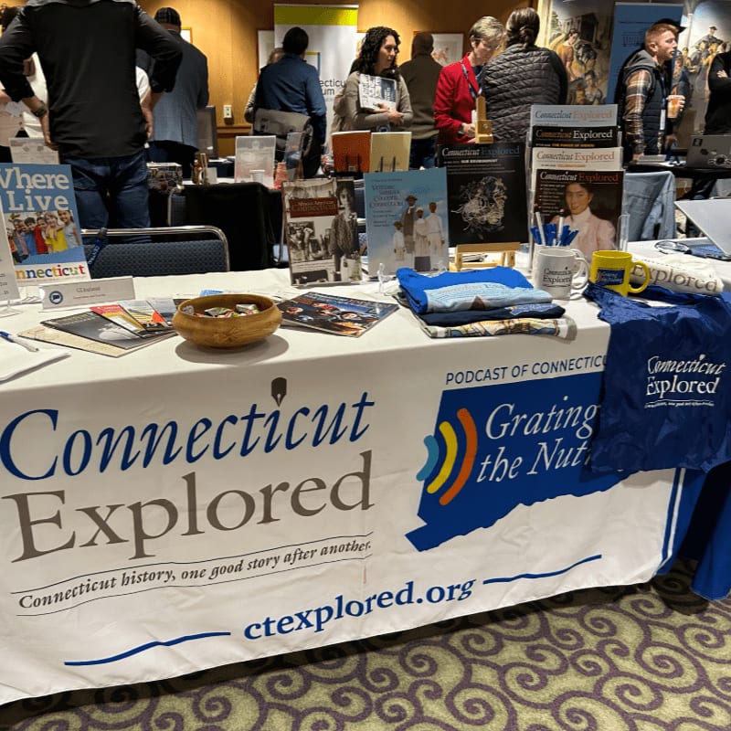 A table covered in a "Connecticut Explored" branded cloth displays books and brochures in the foreground, while several people mingle in a blurred background.