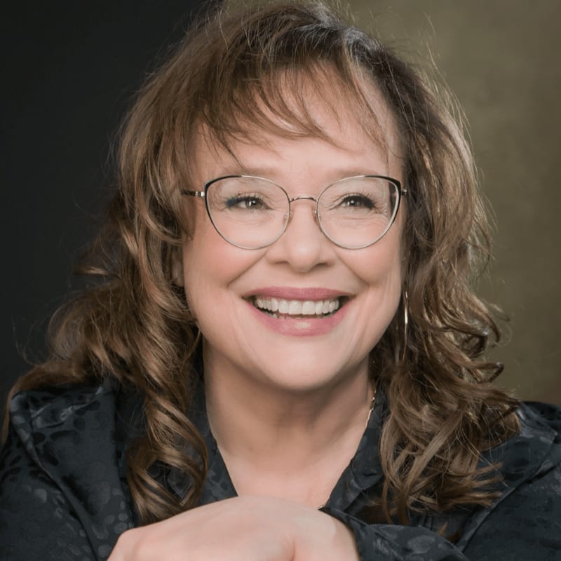 A smiling woman with glasses and wavy brown hair is positioned in the foreground against a blurred, dark neutral background.