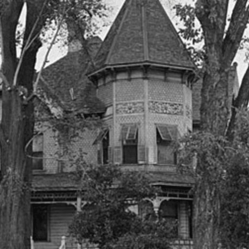Tall trees frame a Victorian house with a prominent turret and decorative trim.