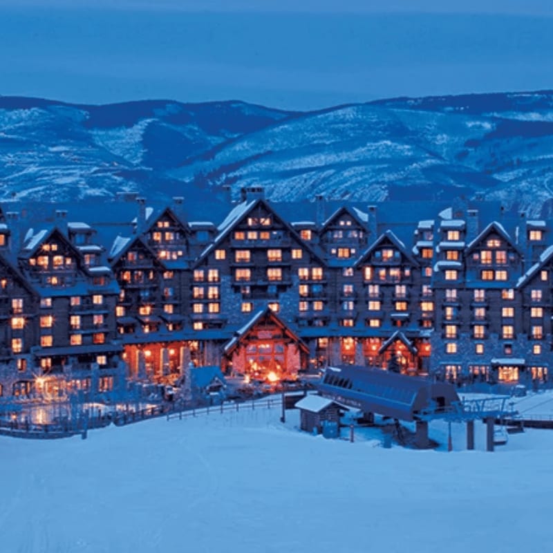 A large, illuminated mountain lodge sits in a snowy foreground against a backdrop of blue, snow-covered mountains.