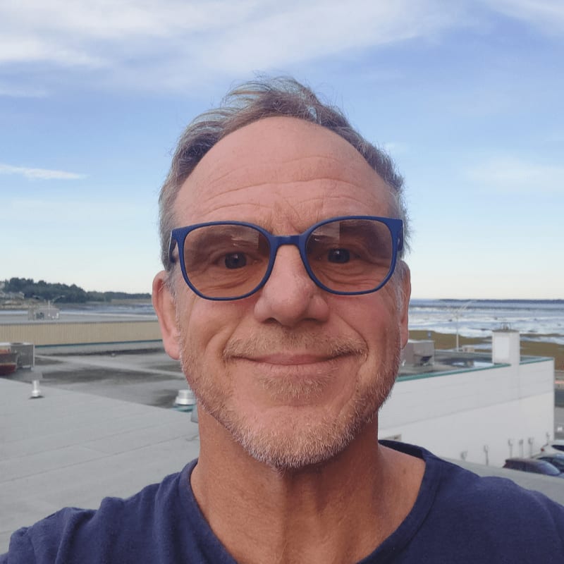 A man wearing blue-rimmed glasses smiles in the foreground against a background of a flat rooftop and a distant coastline under a blue sky.