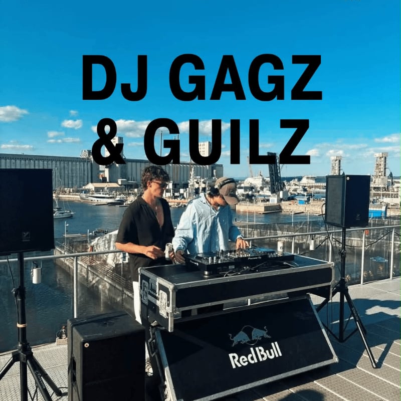 Two DJs perform at a turntable setup on a rooftop overlooking a harbor with ships and industrial buildings under a clear blue sky.