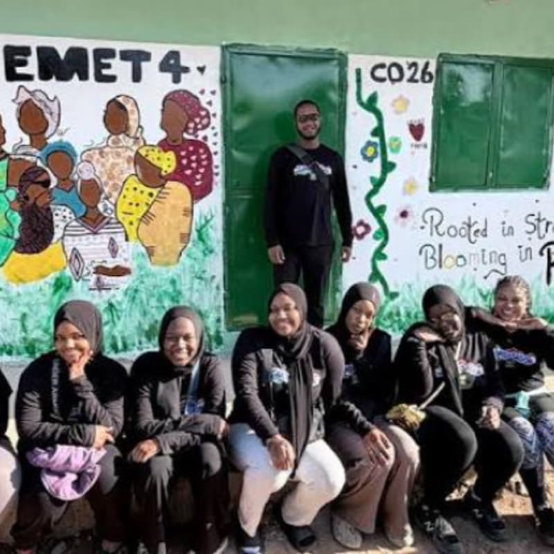 A group of people sit and stand in the foreground in front of a green wall featuring a colorful mural and a closed door.
