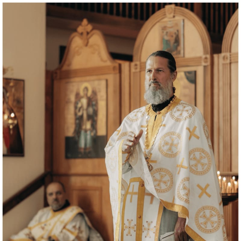 A bearded priest in ornate white and gold vestments stands in the foreground, with another priest and religious icons in a wooden church setting behind him.