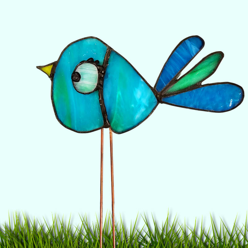A blue stained-glass bird stands on thin wire legs atop a patch of green grass against a pale blue background.