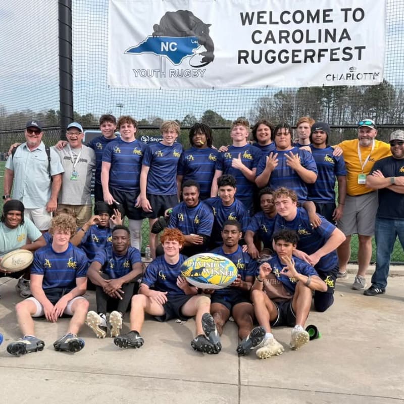 A rugby team poses for a group photo in the foreground beneath a "Welcome to Carolina Ruggerfest" banner in the background.