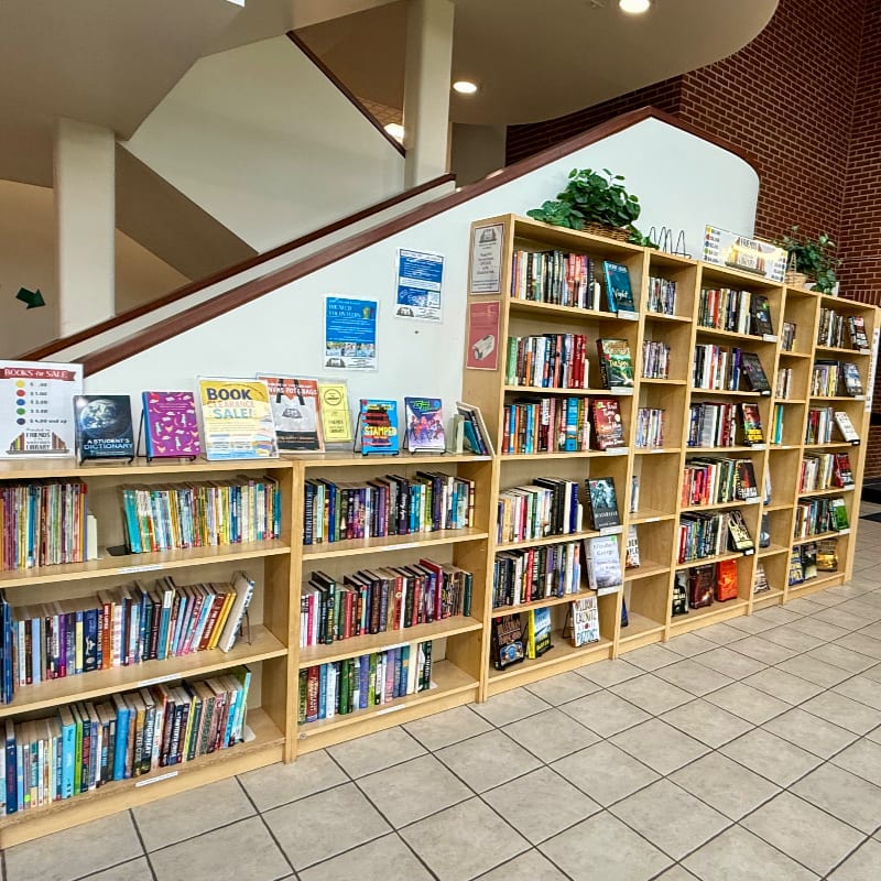 Wooden bookshelves filled with books stand in the foreground against a background featuring a white wall, a staircase, and a brick wall.