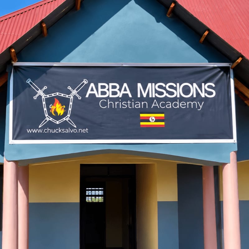 A black banner for Abba Missions Christian Academy hangs in the foreground against a blue building with a red roof in the background.