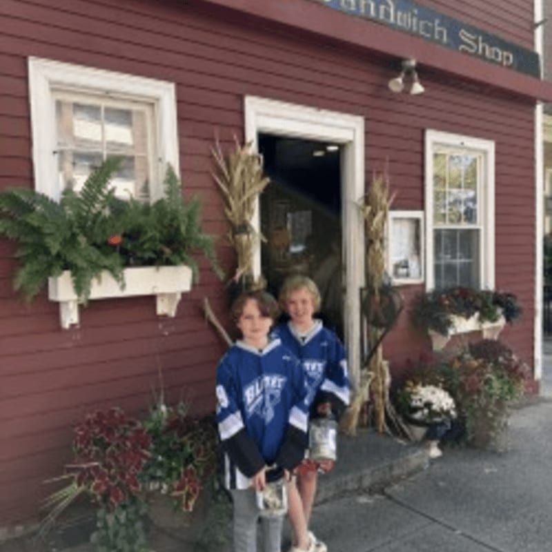 Two young boys in blue hockey jerseys stand in the foreground in front of a red wooden sandwich shop decorated with autumn plants.