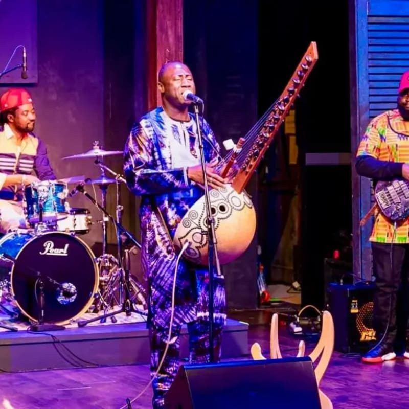 A musician in a blue patterned outfit plays a kora in the foreground, with a drummer and another musician performing on a stage in the background.