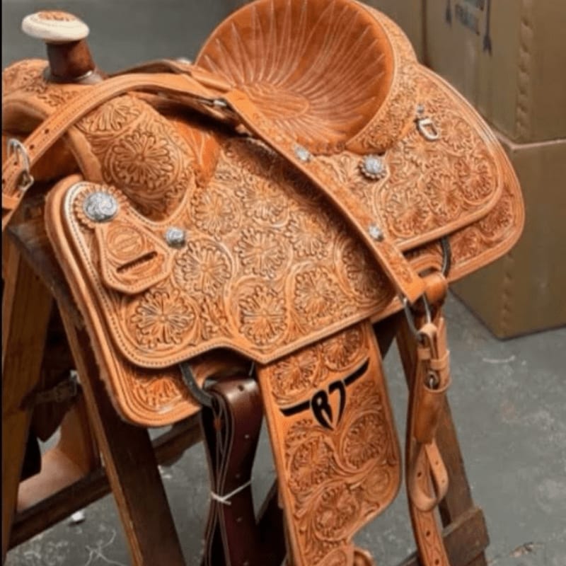 An intricately carved brown leather saddle rests on a wooden stand against a backdrop of cardboard boxes.