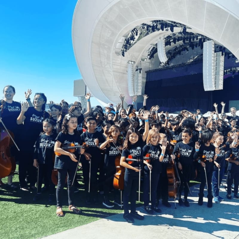 A large group of children holding string instruments stand together in the foreground beneath a massive, white curved outdoor stage structure.