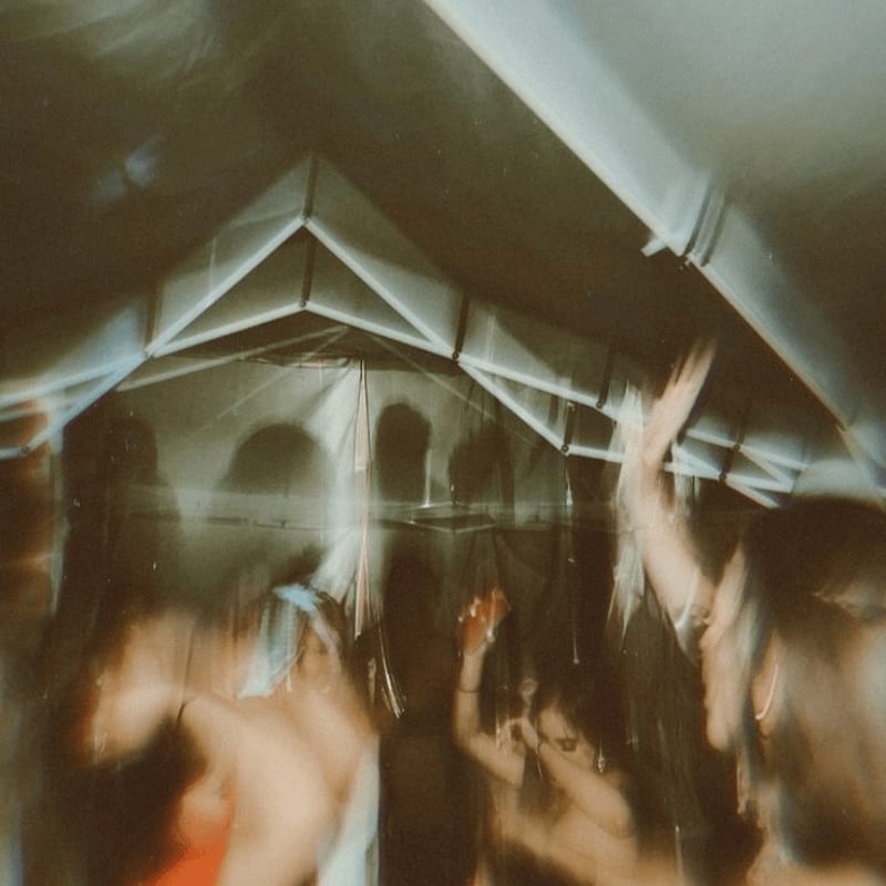 Blurred figures dance in the foreground beneath the peaked ceiling of a dimly lit tent.