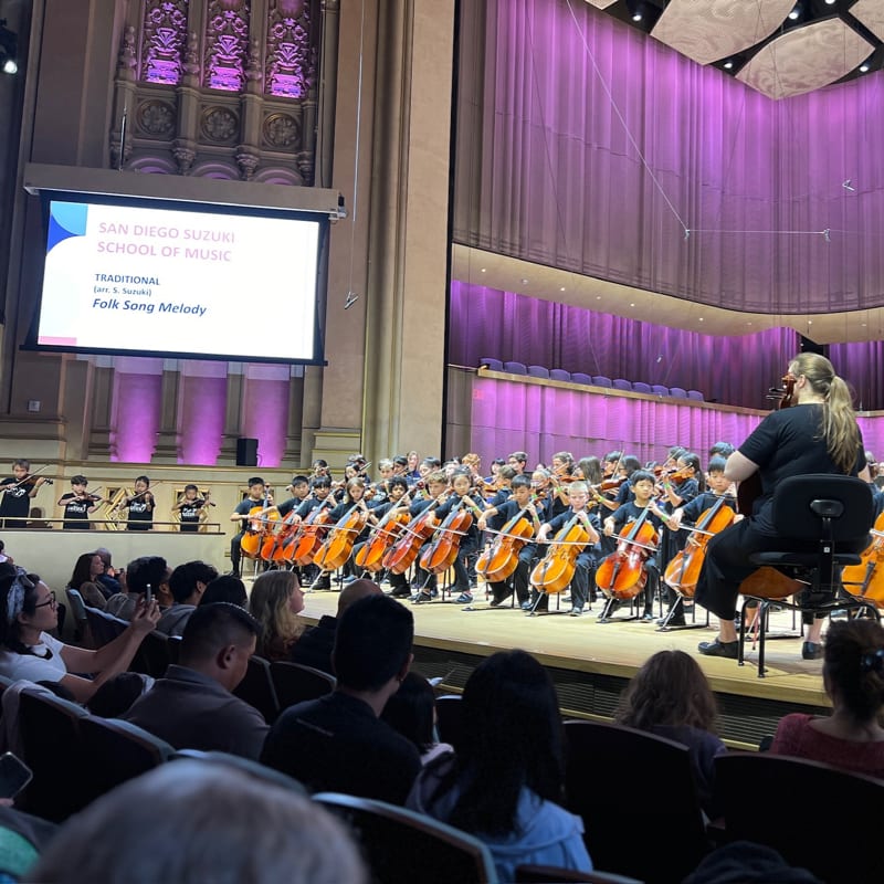 An orchestra of young cellists performs on a stage in front of an audience, with a large screen and purple-lit architectural details in the background.