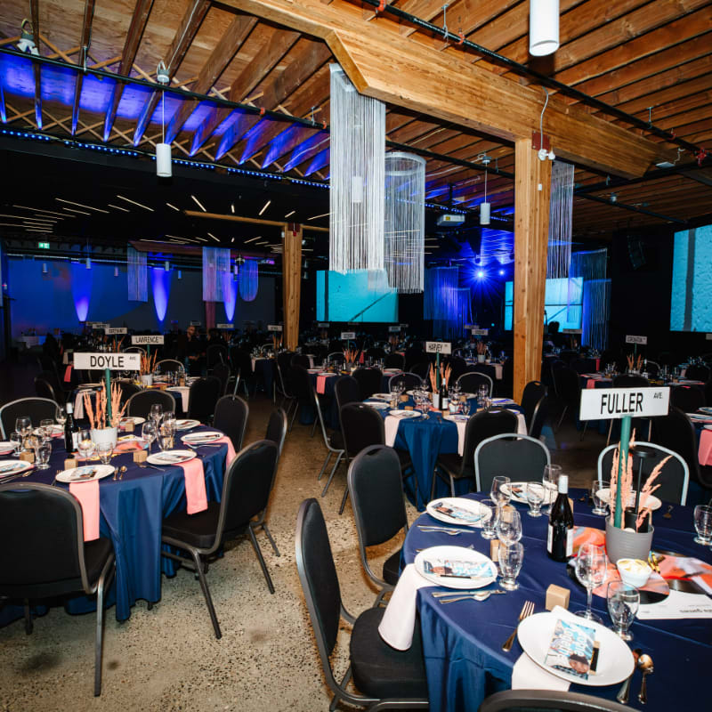Round tables with blue cloths and place settings occupy the foreground of a rustic event space with wooden beams and blue ambient lighting in the background.
